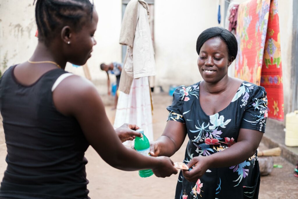 A woman in Uganda smiles as she takes payment from another woman for liquid soap. They are in a courtyard together.