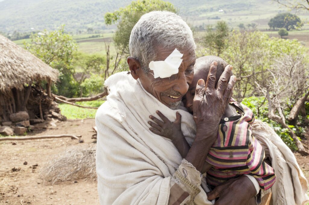 Ethiopian grandfather Mekonen with a white eye bandage after his cataract surgery happily carries his grandchild