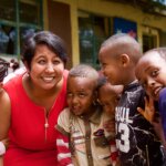 Nafisa Baboo smiling with a group of 6 Ethiopian children. Nafisa has lightbrown skin, short dark hear and wears a red sleaveless dress.