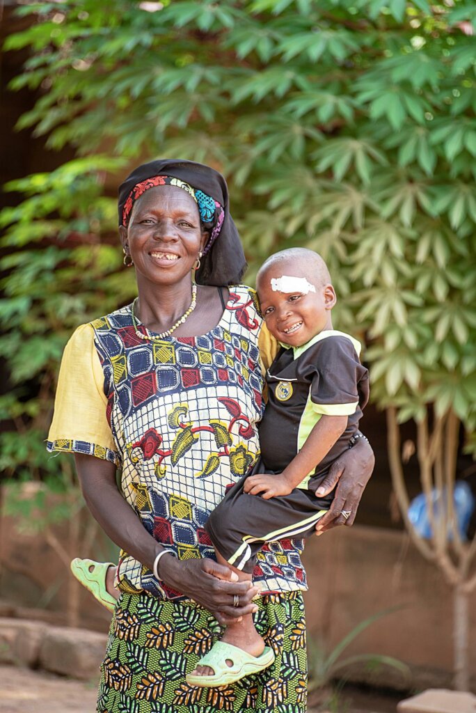 young Burkinabe boy Zakaria with his mother after his eye surgery. both are smiling and Zakaria still wears his white eye bandage.