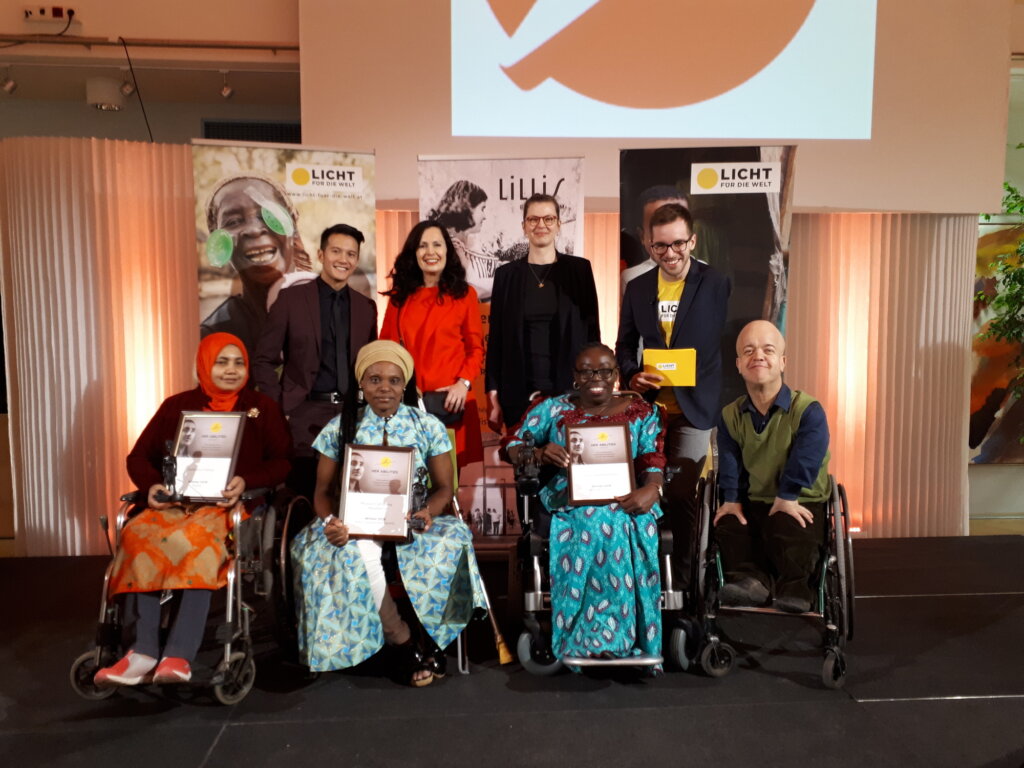 Eight people smile, looking at the camera. They are on a stage with a background with Light for the World banners. The photo shows the Award ceremony for the 2018 Light for the World Her Abilities Award Laureates.