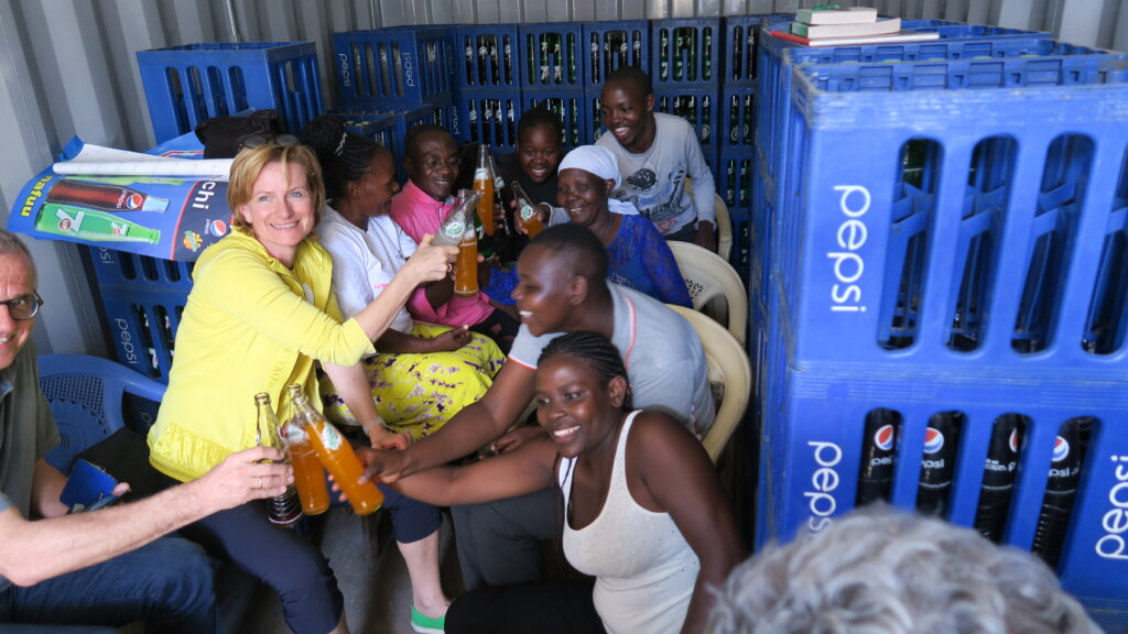 Several people, including Karin Krobath to the left, clink/'cheers' soft drink bottles. They are surrounded by blue boxes. Many are participants of the Light for the World InBusiness Initiative