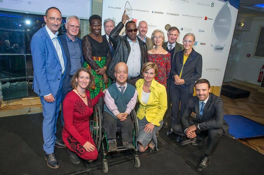Karin Krobath, dressed in a yellow jacket, kneels and smiles at the camera. She is pictured alongside several other members of the International Board of Light for the World including Tom Shakespeare, to her left, and Woldesenbet Brhanemesqel, who stands behind Karin holding a trophy in his hands/on his head.