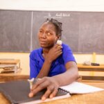 Nadège sitting in class using an audio player and her Braille slate. She is a Burkinabe teen wearinga blue T-shirt and her hair in braids