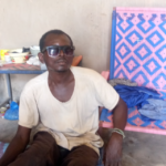 Jean-Baptiste, wearing dark sunglasses, sits on a mat in his house, leaning against one of his hand-woven chair