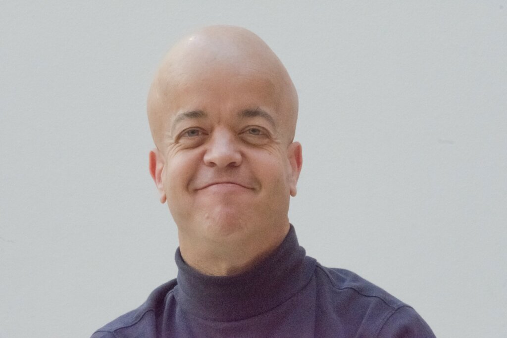 Portrait picture of man smiling. He wears a dark blue polar neck jumper. He looks straight at the camera.