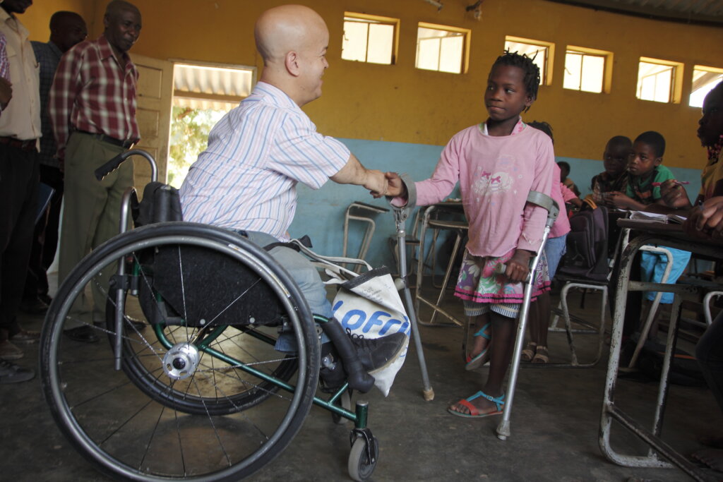 White man in wheelchair shakes hand with young girl from Mozambique who uses crutches. They are in a brightly coloured yellow and blue school classroom.