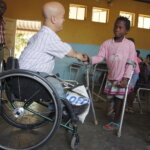 White man in wheelchair shakes hand with young girl from Mozambique who uses crutches. They are in a brightly coloured yellow and blue school classroom.