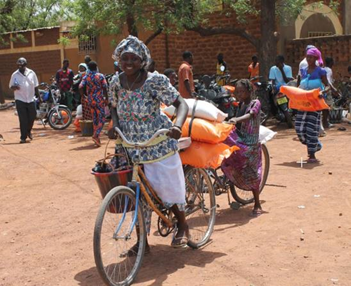 Two Burkinabe girls in identical skirts stand behind bags of food, buckets and school supplies. In the background are many motorcycles and several internally displaced people with bicycles. photo credit: KIO photography 