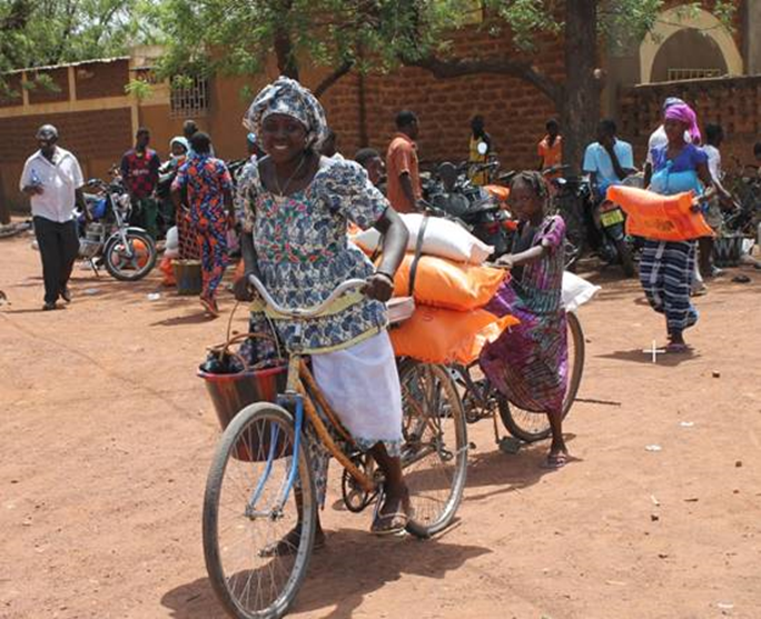 Two Burkinabe girls in identical skirts stand behind bags of food, buckets and school supplies. In the background are many motorcycles and several internally displaced people with bicycles. photo credit: KIO photography