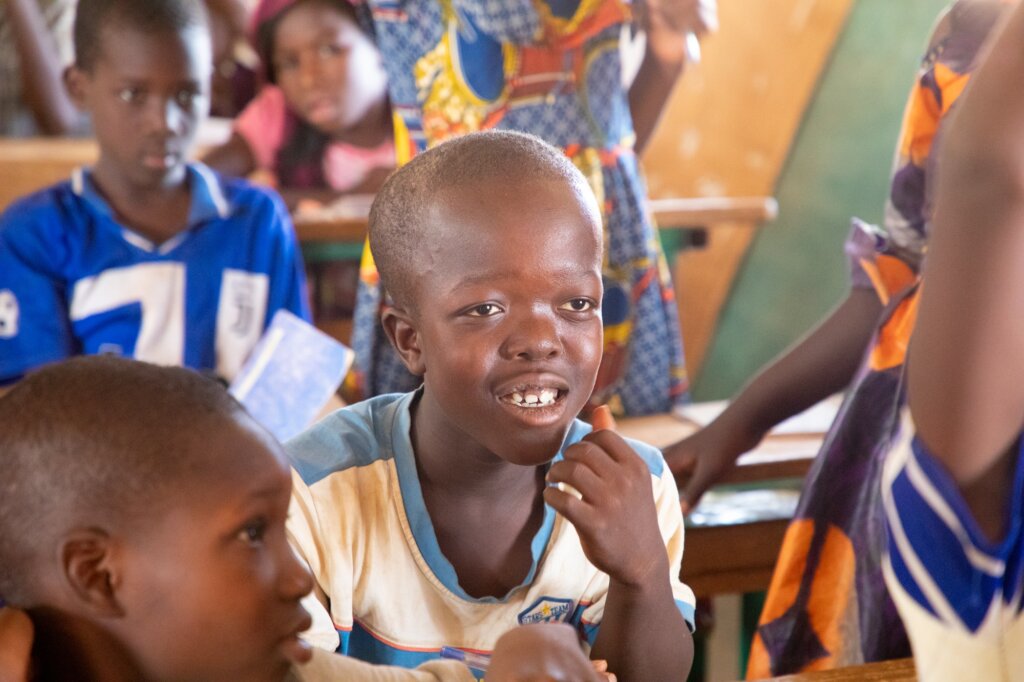 12-year-old Burkinabé boy sits in class with a smile, surrounded by his friends. Alira is of small stature and has a high forehead.