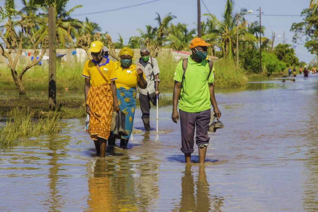 4 Mozambican adults wade through a flooded street. At the rear is an older man with one leg and crutches.