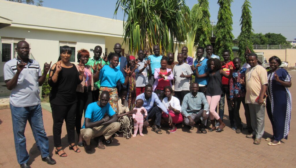 a group of about 20 South Sudanese members of the disability rights community. They pose for a group photo in front of a low building and make signs with their hands.