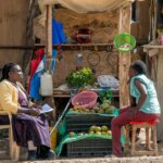two Kenyan women seated in front of a fruit and vegetable stall. the woman on the left wears orthopaedic shoes and holds a notepad in her hands as she interviews the other woman about the impact of COVID