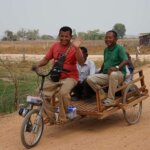 four Cambodian project participants riding a transport tricycle on a dirt road. The driver is male and waves and smiles. 2 men and one woman sit behind him on the cart.
