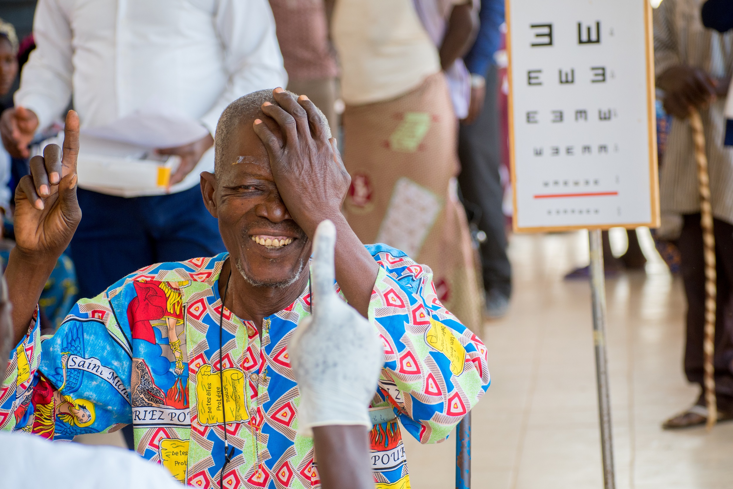 Paul, an older Burkinabe man with short gray hair and beard in a colourful cotton short, covers one eye and smiles as he does a vision test after his cataract surgery. behind him are blurred silhouettes of people and a vision board.