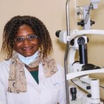 Burkinabe woman with glasses and shoulder-length curly hair in a white lab coat and a facemask under her chin standing next to ophthalmic machine