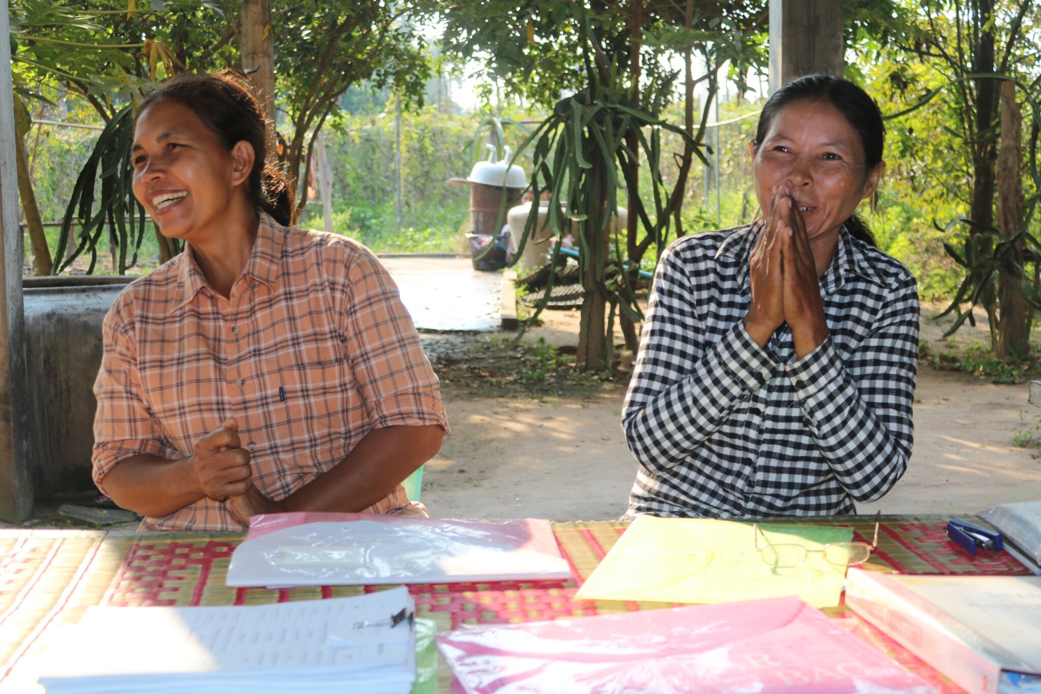 Two Cambodian women participate in a workshop for women with disabilities in Pursat. They sit outside behind a table with documents. Both are laughing. The woman on the right has her hands, palms pressed together, up in fron of her mouth.