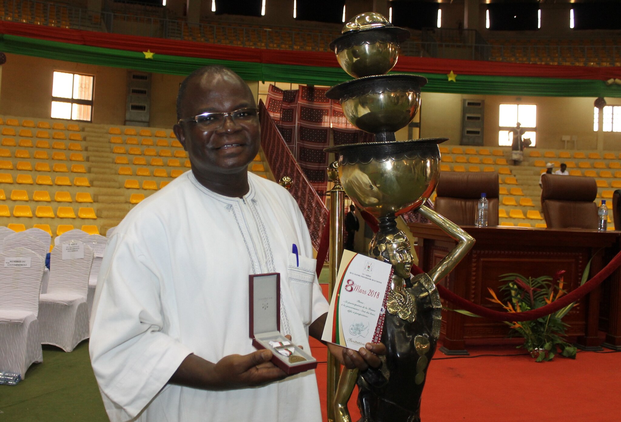 Elie Bagbila a 58-year-old Burkinabe man with glasses and a white traditional shirt stands in an auditorium holding a medal of distinction. next to him is a bronze sculpture taller than him.