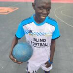 Jimmy ist Teil des Blindenfußballteams im Südsudan. Er trägt ein weißblaues Trikot, hält den Ball in seinem linken Arm und lächelt in die Kamera. Hinter ihm ist das Fußballfeld zu sehen.