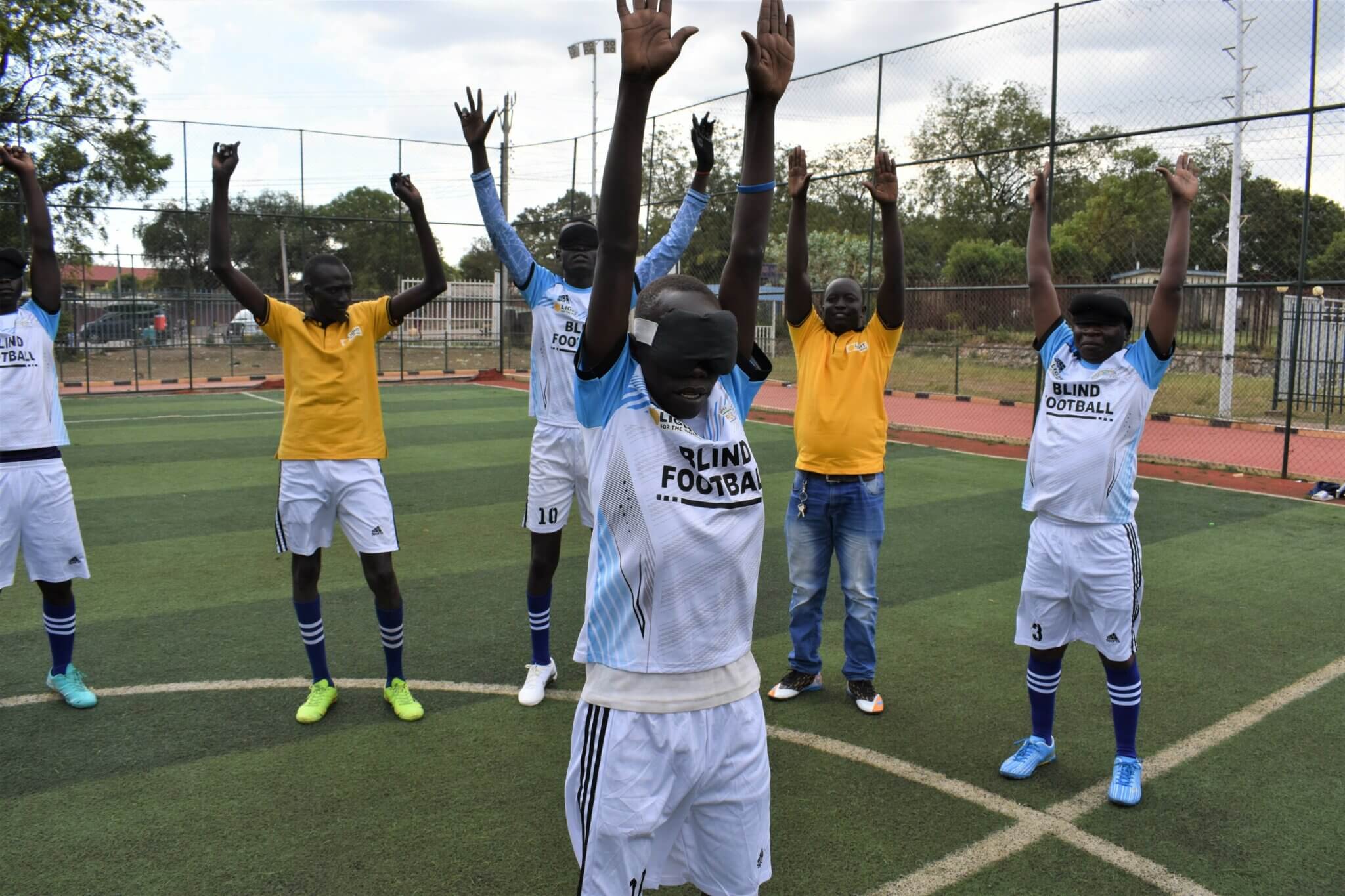The members of the blind football team stand together in a circle on the pitch and stretch their hands in the air. They are warming up.