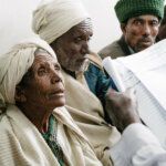 An older Ethiopian woman wearing white traditional clothing and headscarf sits next to three men in the waiting room of a mobile eye health outreach. She responds to questions from a health professional in a white coat.