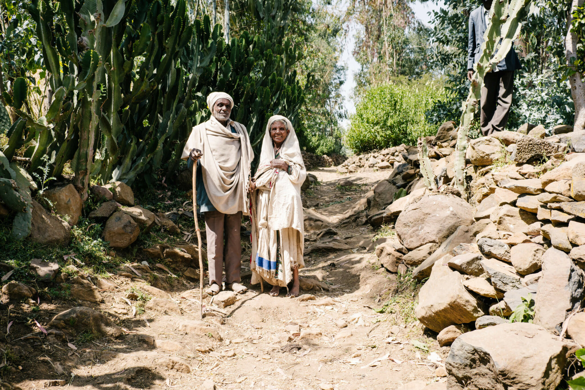 Mullu, an older Ethiopian woman, and her husband, both in traditional white cotton clothes and head covers, on a dirt path framed by rocks.