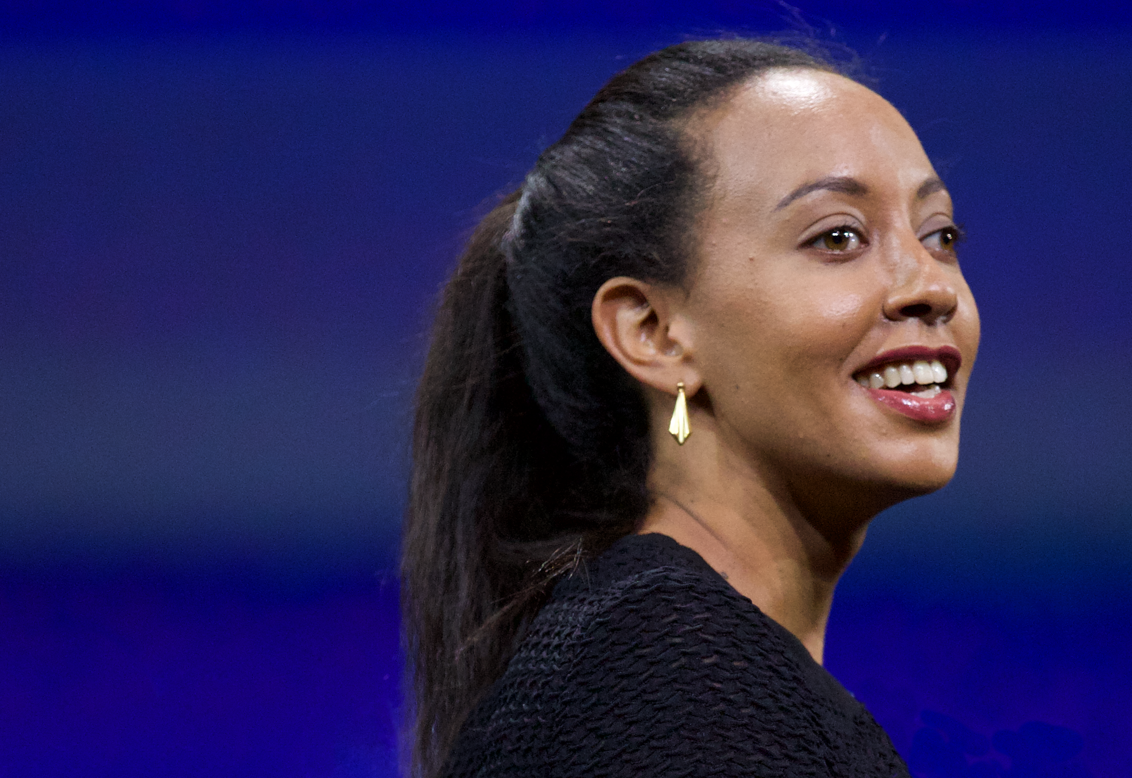Profile picture of Haben Girma. Plain blue background headshot. Haben is smiling, wearing navy blue cardigan, gold earrings, hair tied back. Head turned to the right.