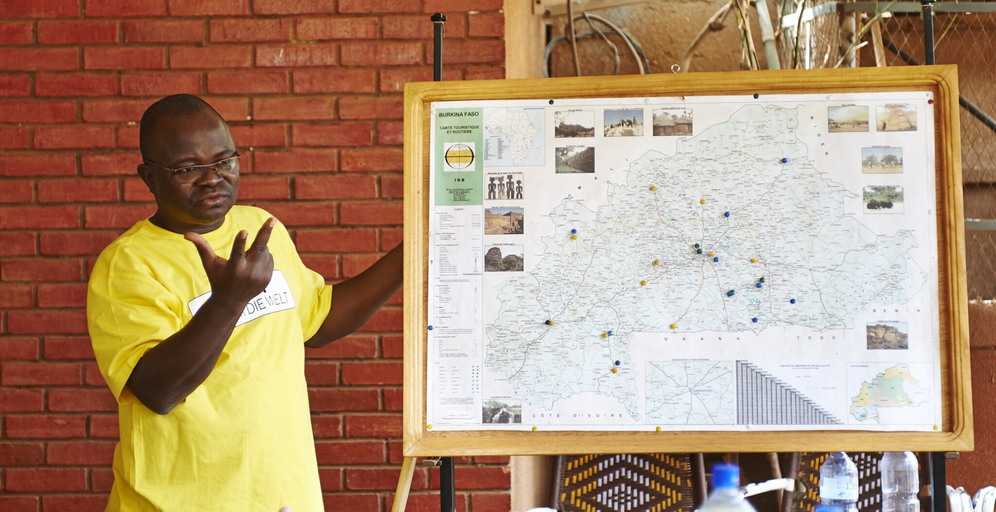 Elie Bagbila, a Burkinabe man with glasses and a yellow T-shirt speaks next to a Burkina Faso map