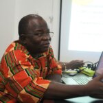 Elie Bagbila, a Burkinabe man in his mid-50ies with glasses and an orange patterned shirt gives a presentation