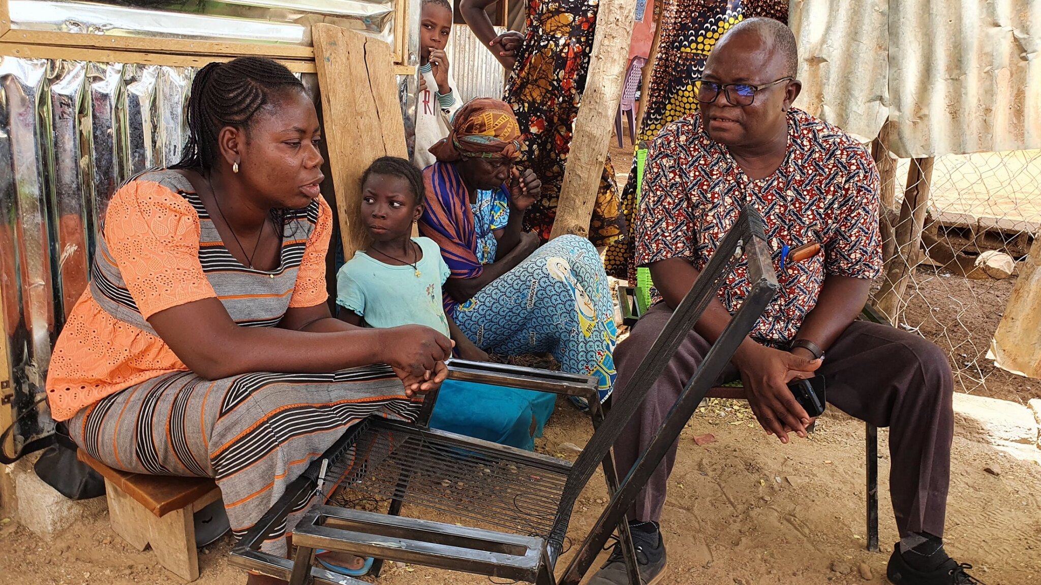 a young Burkinabe woman is repairing a chair, next to her sits a middle-aged black man with glasses and children sit in the background.