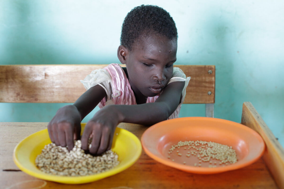 Amandine has her hands in a yellow plate and feels beans and millet in it. Next to it is an orange plate into which she throws the small millet grains after she has been able to feel them.