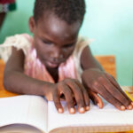 Amandine aus Burkina Faso sitzt an einem Tisch in der Inklusionsschule in Kaya, Burkina Faso. Ihre Hände liegen auf weißem Papier und sie übt, die Brailleschrift zu lesen. Sie wirkt sehr konzentriert.