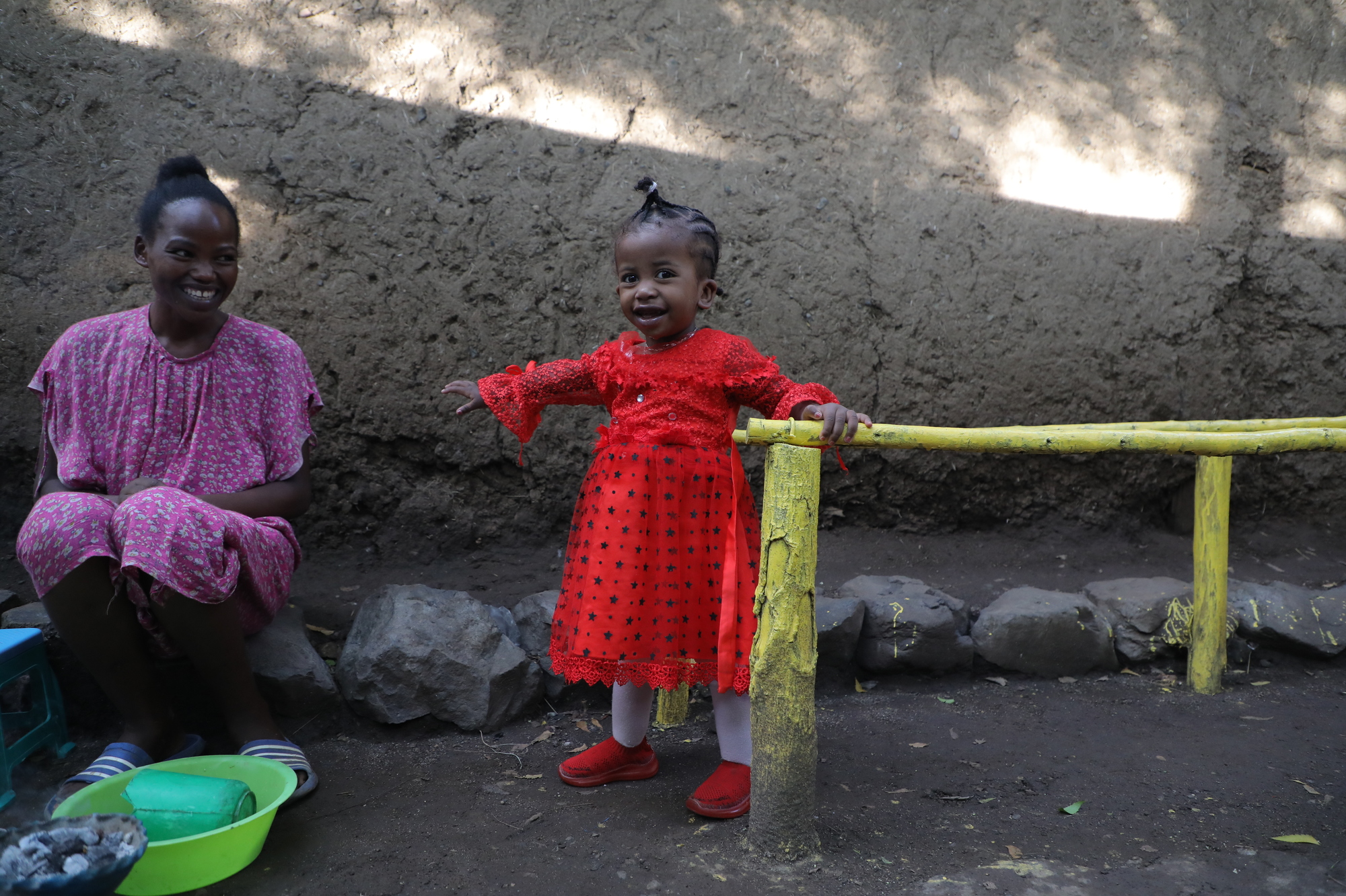 A small girl in a red dress stands smiling next to walking bars and smiles into the camera. Her mother sits next to her in a violet dress and smiles at her daughter.