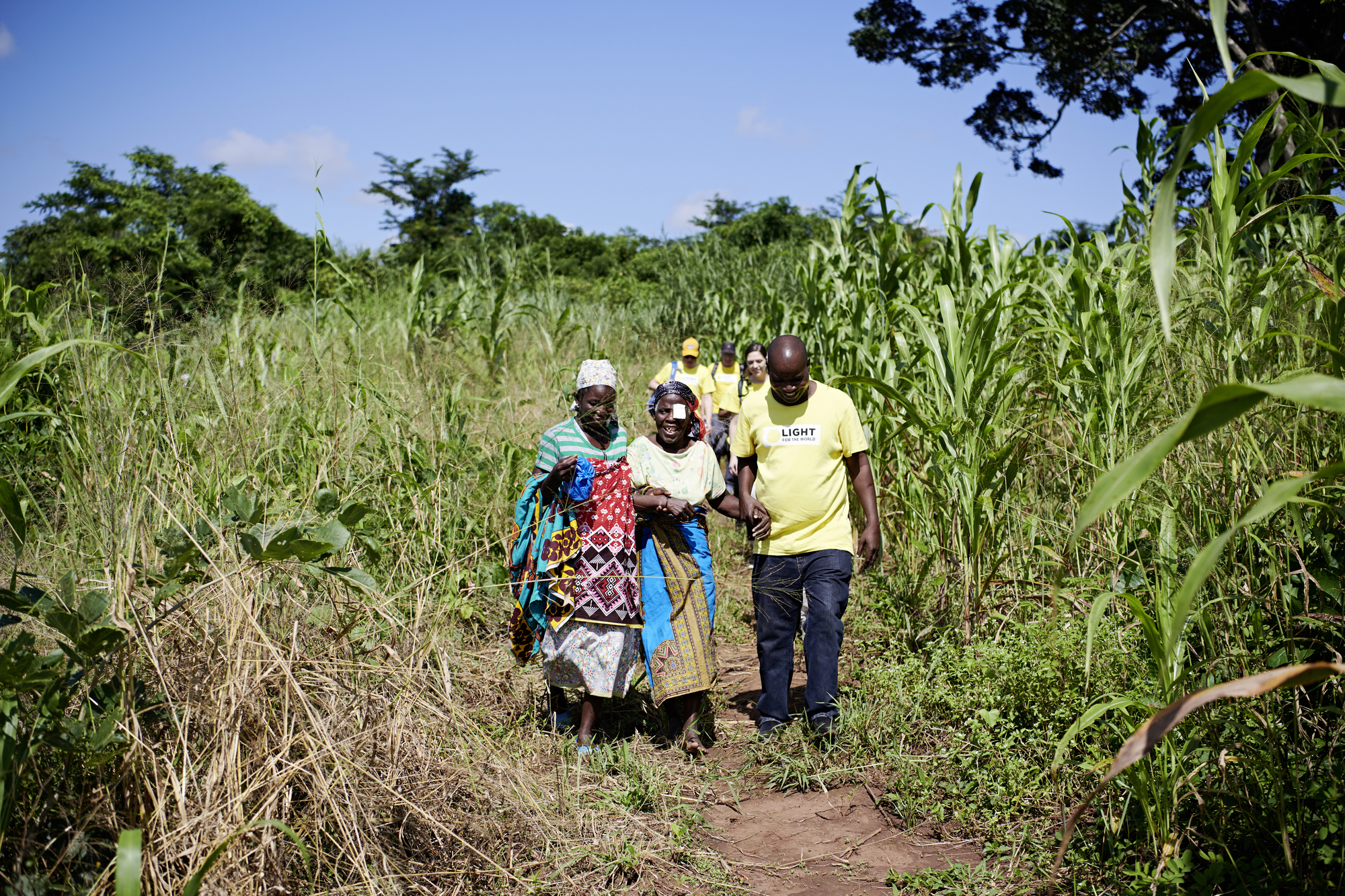 A slideshow image shows a woman being escorted back home after an operation. Next to her walks her relative and an aid worker from Light for the World in Mozambique. They accompany her through the fields back home.