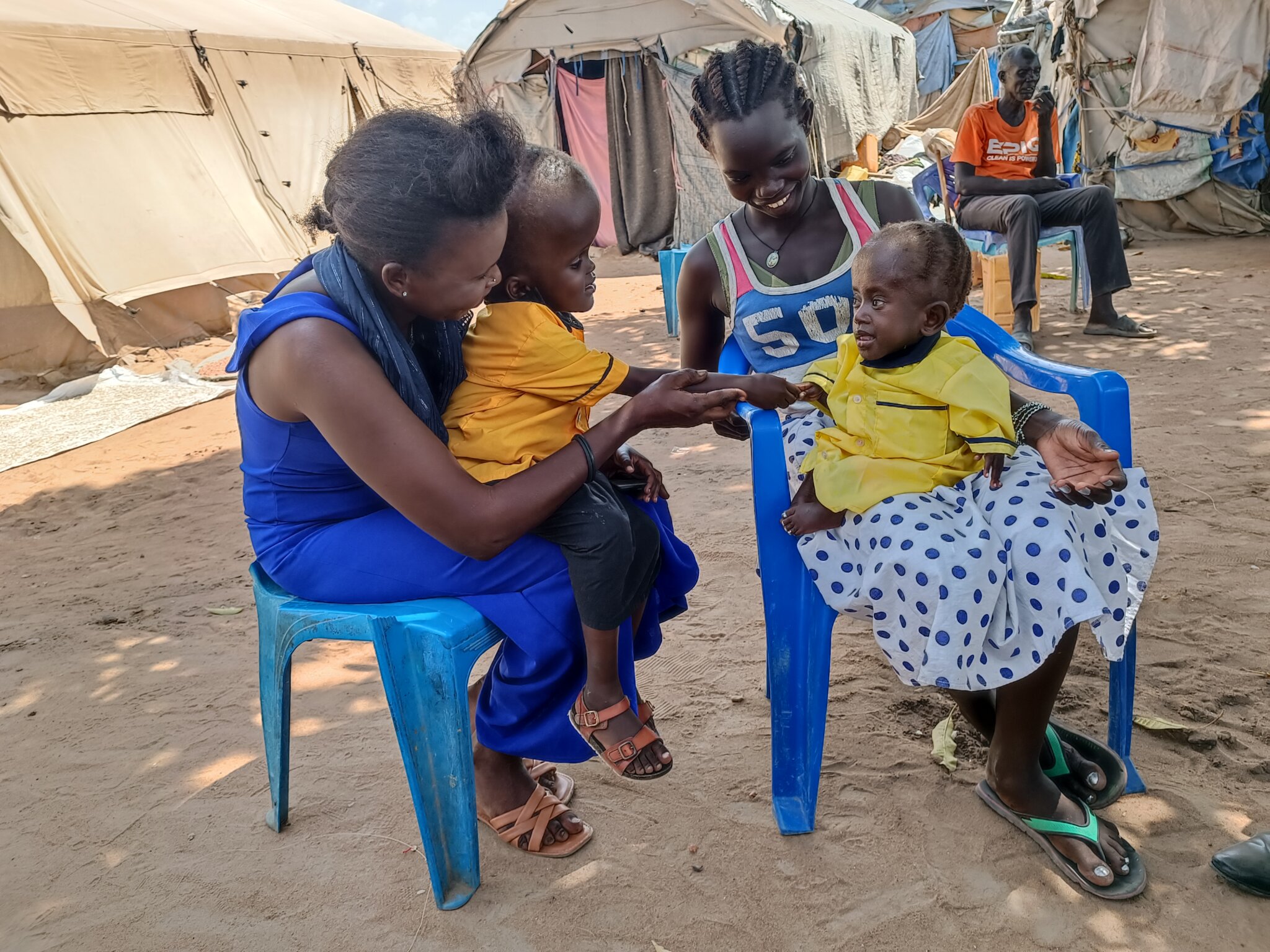 To women sit on blue chairs with children in their lap. The children reach for each other’s hands and the mothers are happily smiling.