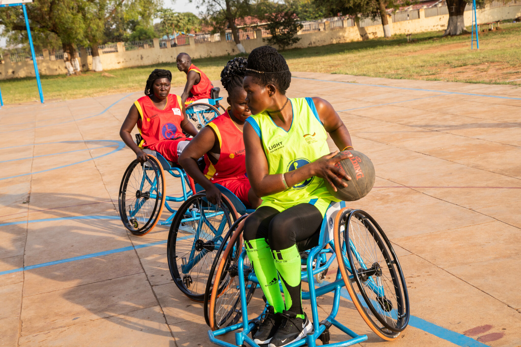 Four women in wheelchairs are playing basketball on a big field. One woman in a yellow shirt is dribbling the ball.
