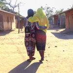 Hero image of two women in Mozambique walking with their backs to the camera along a lane between houses.