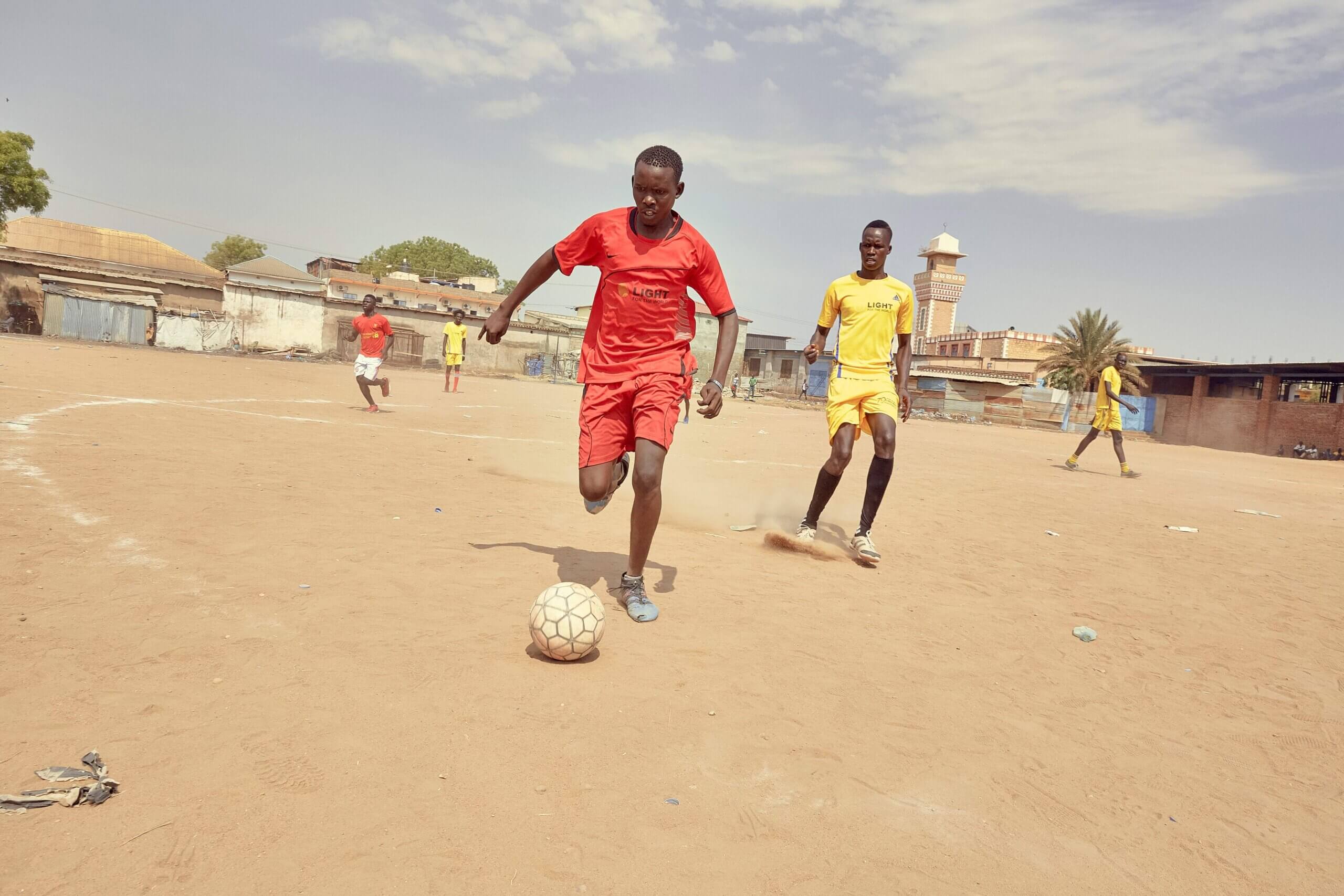 Two members of the Sports for Peace team, one in red and one in yellow, play football on a dusty pitch in South Sudan.