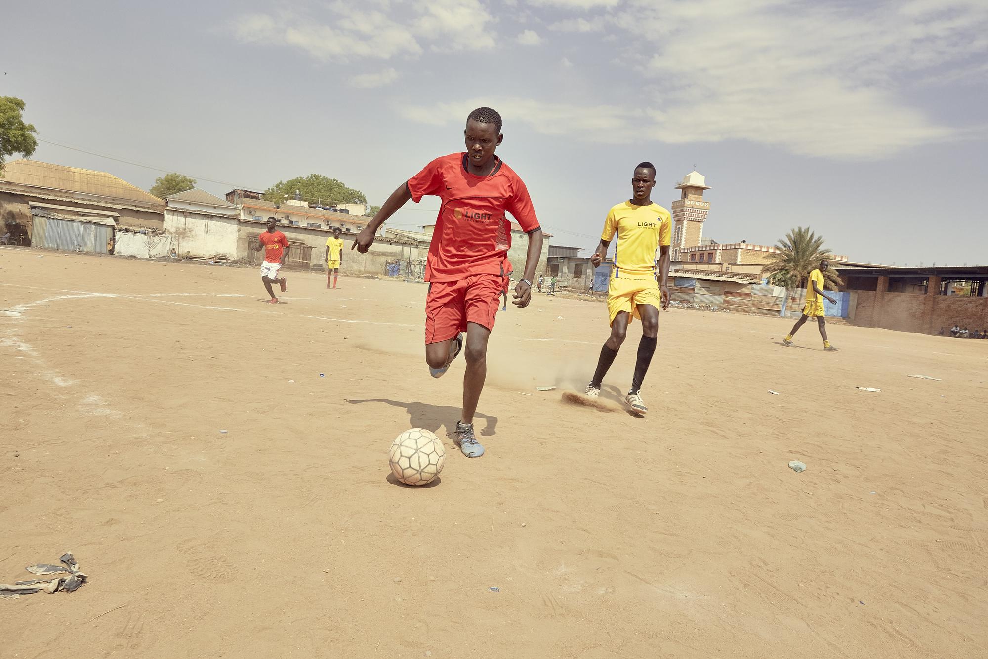 Two members of the Sports for Peace team, one in red and one in yellow, play football on a dusty pitch in South Sudan. 