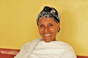 A woman from Ethiopia smiles broadly at the camera. She wears a black and white head wrap and white shawl, and is sitting with her back to a yellow wall.