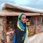 A woman wearing a blue, green and yellow headscarf looks towards the camera while she holds on to the fenced front of the food stall she sells from in South Sudan.