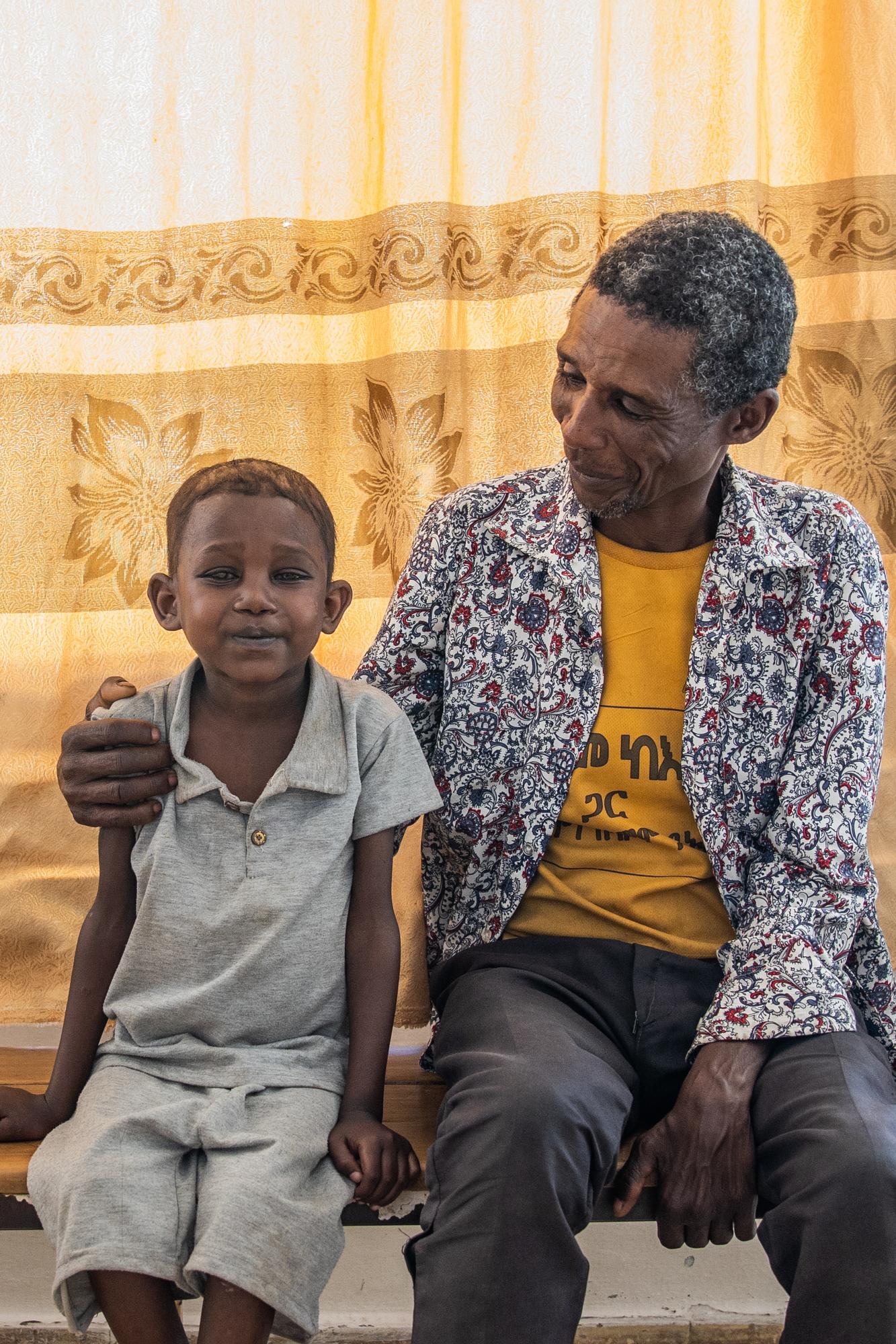 A 10-year-old boy sits next to his smiling dad, in front of a leaf-patterned curtain. His dad has his arm around his shoulder and is smiling down at him.