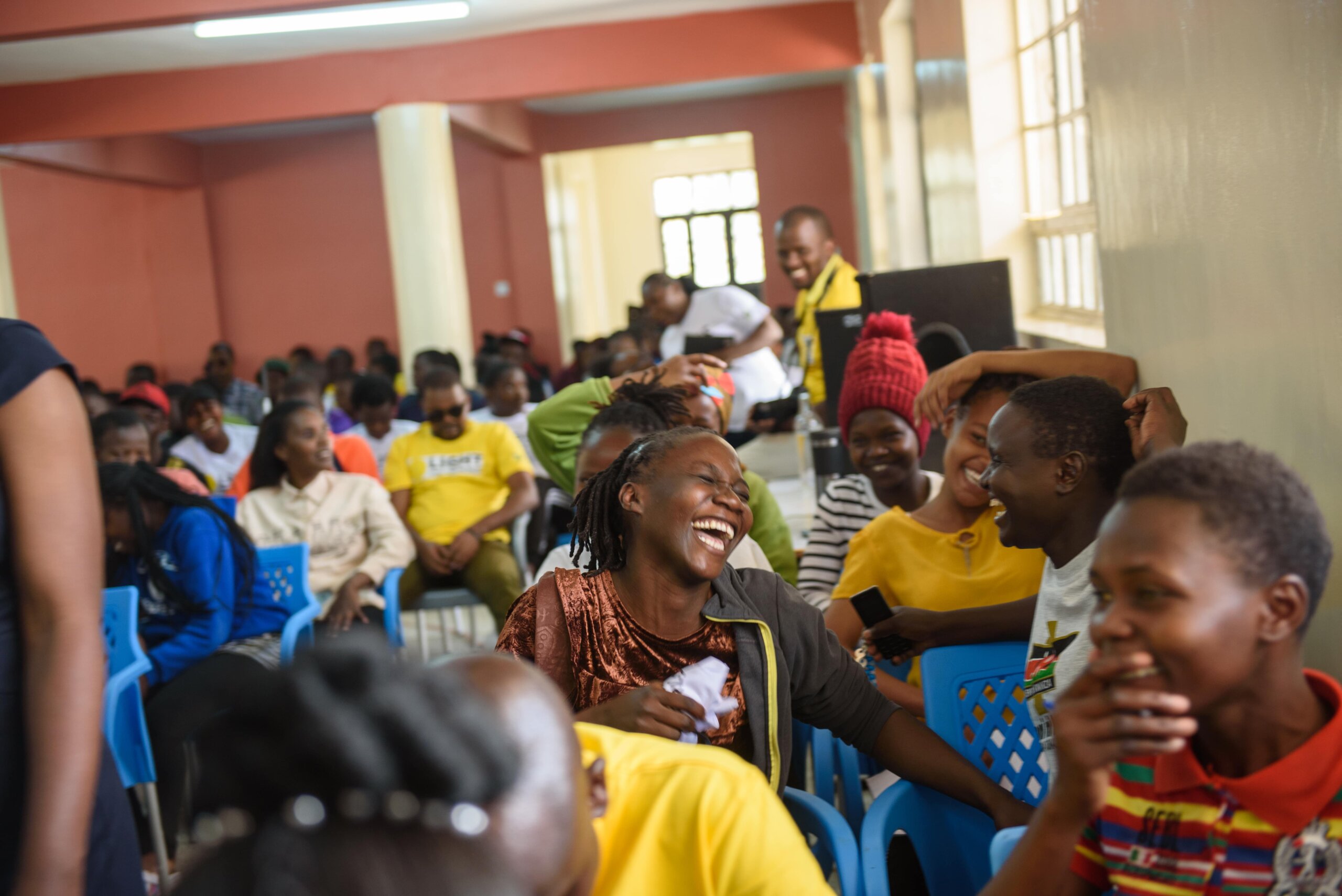 The image shows a room full of people, with a women smiling and laughing in the centre. Many people are smiling and a few are wearing Light for the World t-shirts.