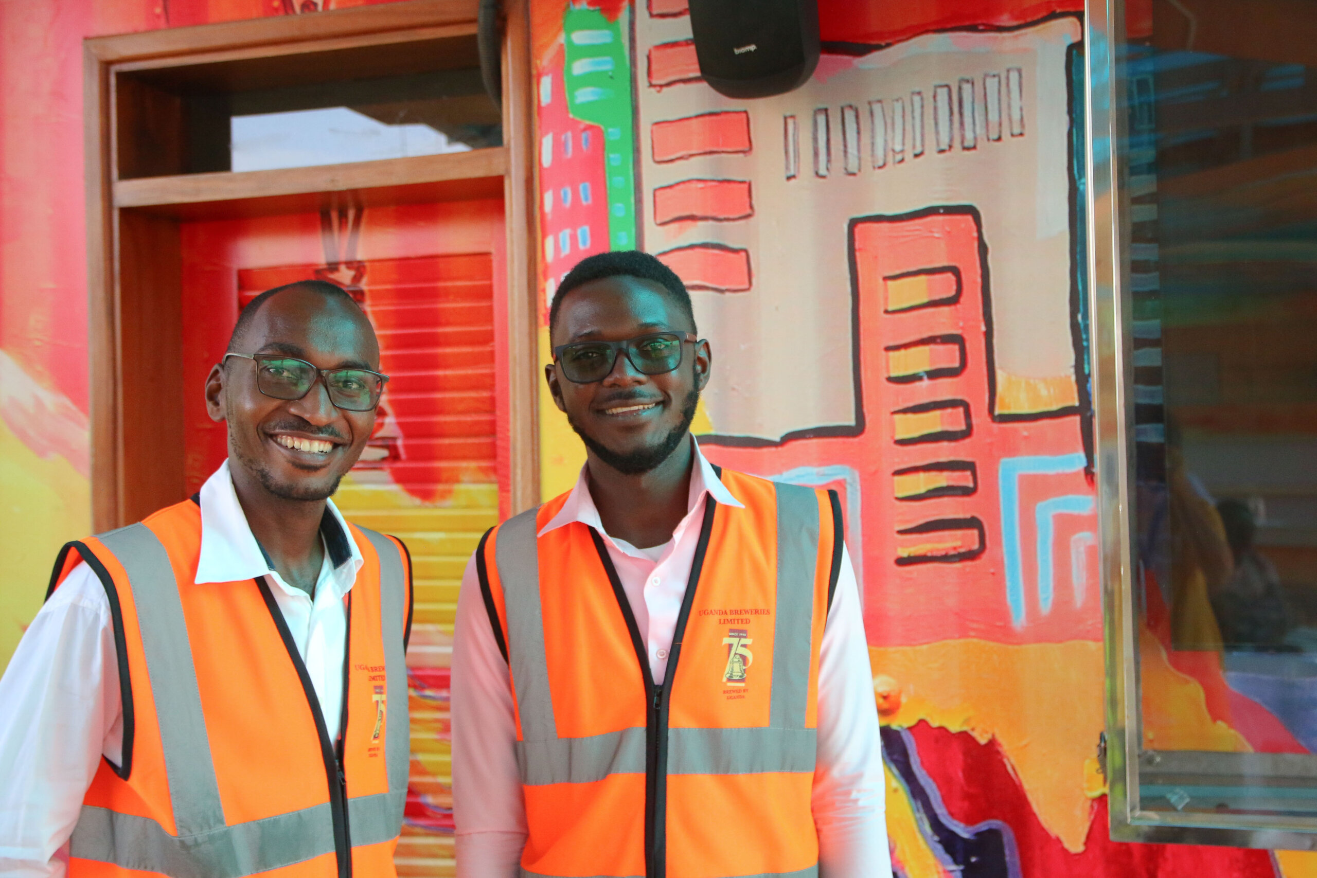 The image shows Higenyi Stuart Cypria (right), pictured smiling next to his team manager, Francis Xavier Kalanzi (left) wearing fluorescent work vests.