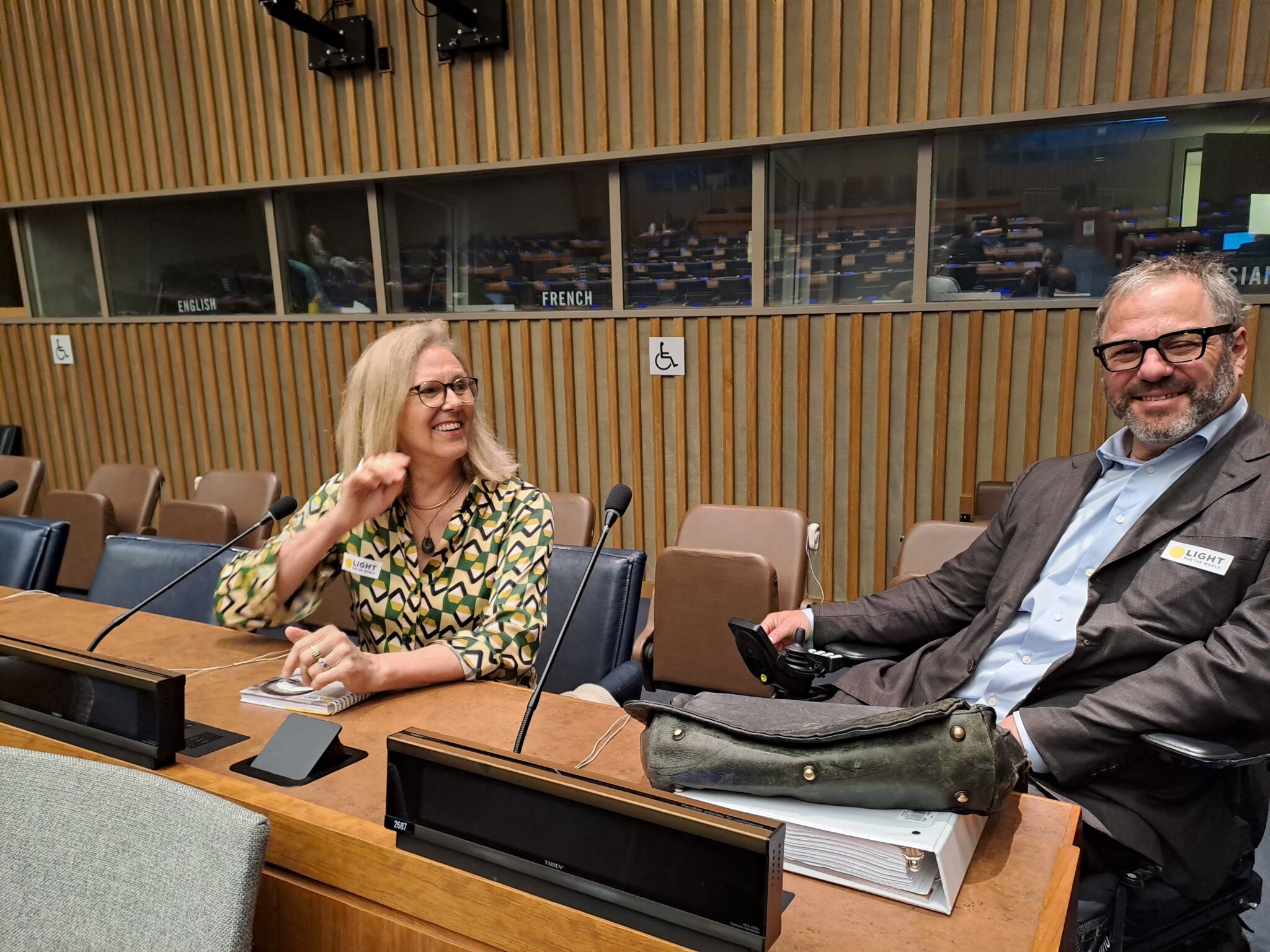 Marion and David laugh together seated inside a UN conference room. There are microphones in front of them, Marion sits on the left, and David on the right.