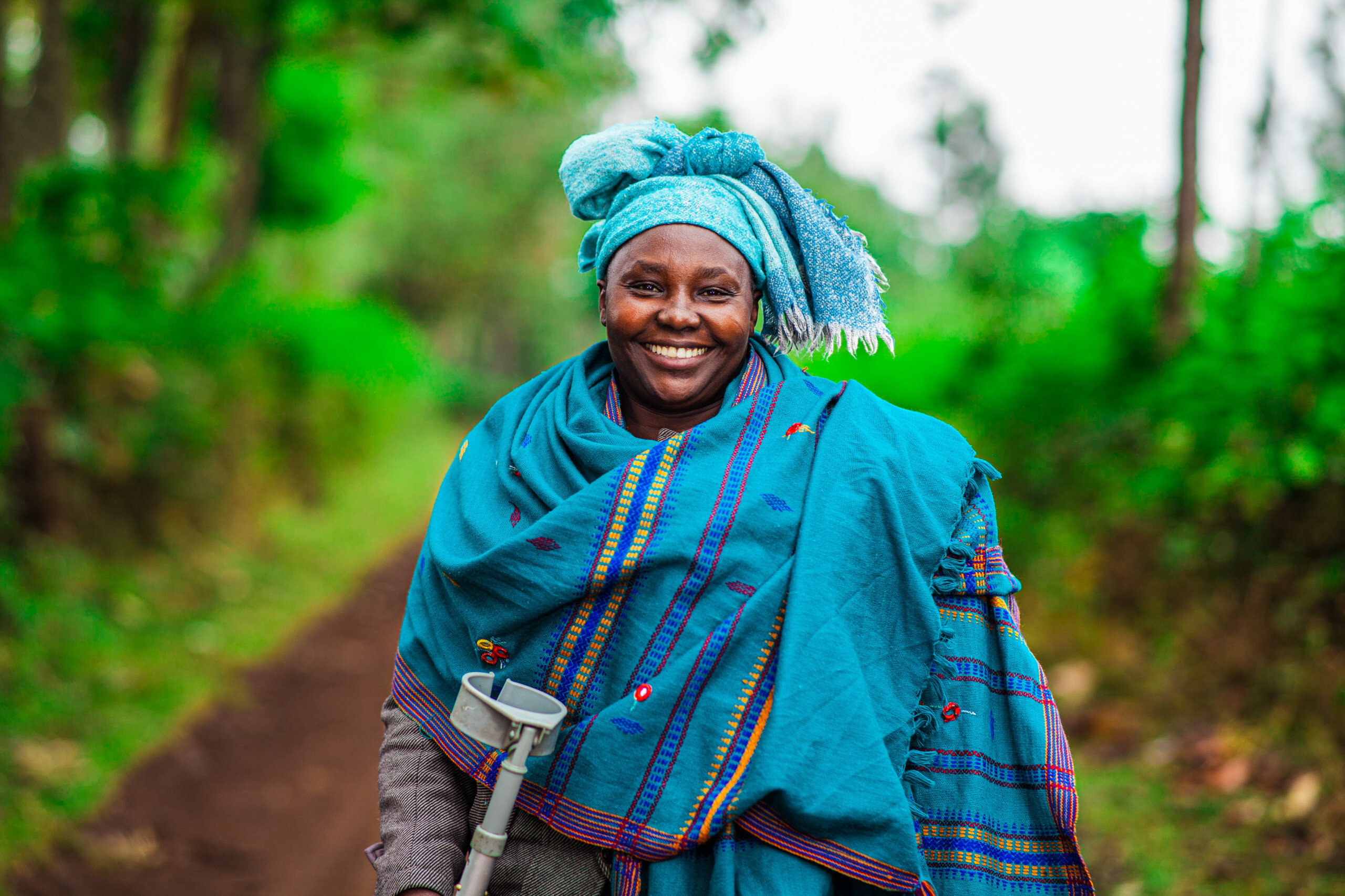 Lucy smiles outside, surrounded by greenery, wearing a patterned blue scarf and holding a mobility aid.