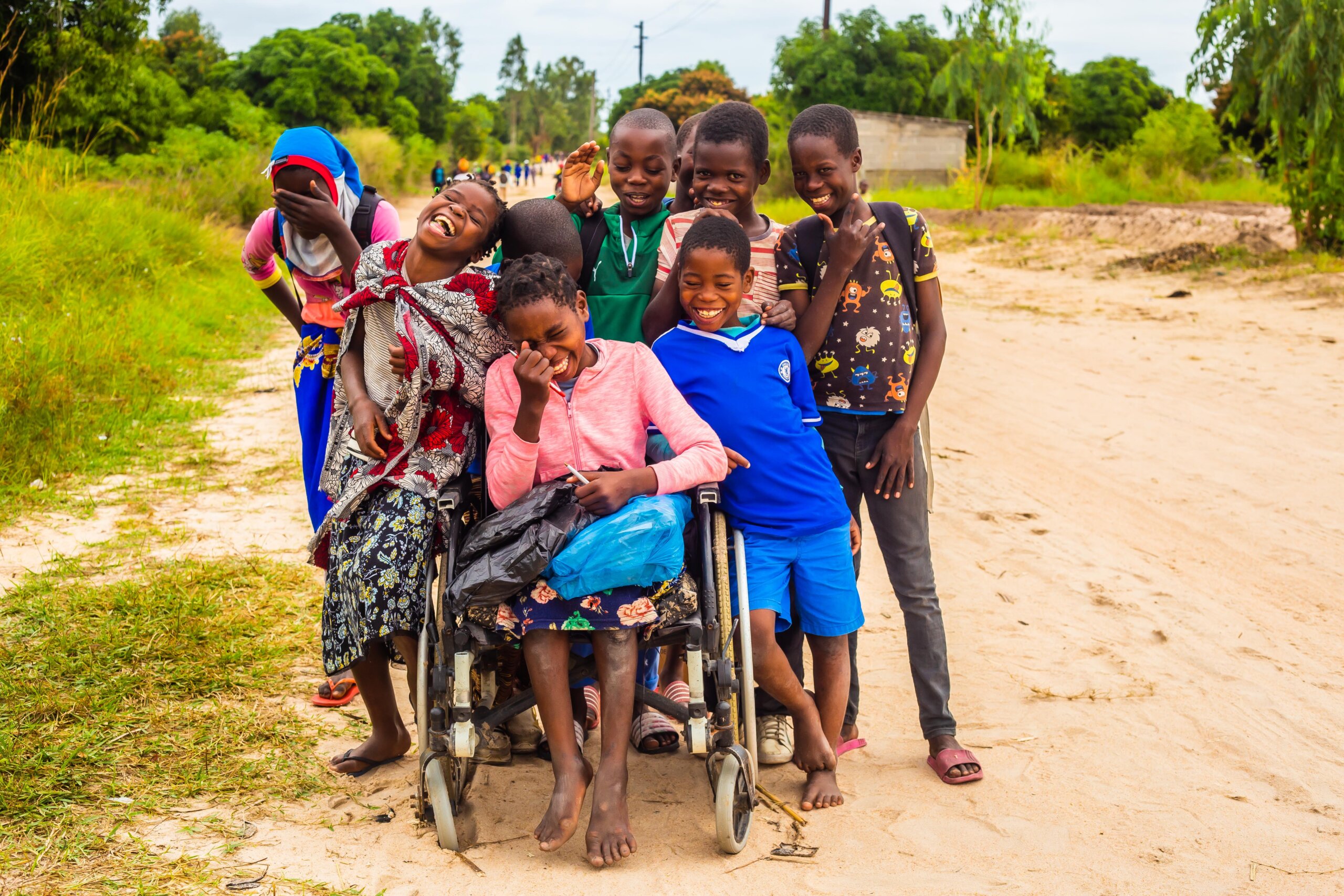 Teresa laughs outside, with 8 of her friends standing behind her. She sits in a wheelchair and holds her schoolbags and pencils.