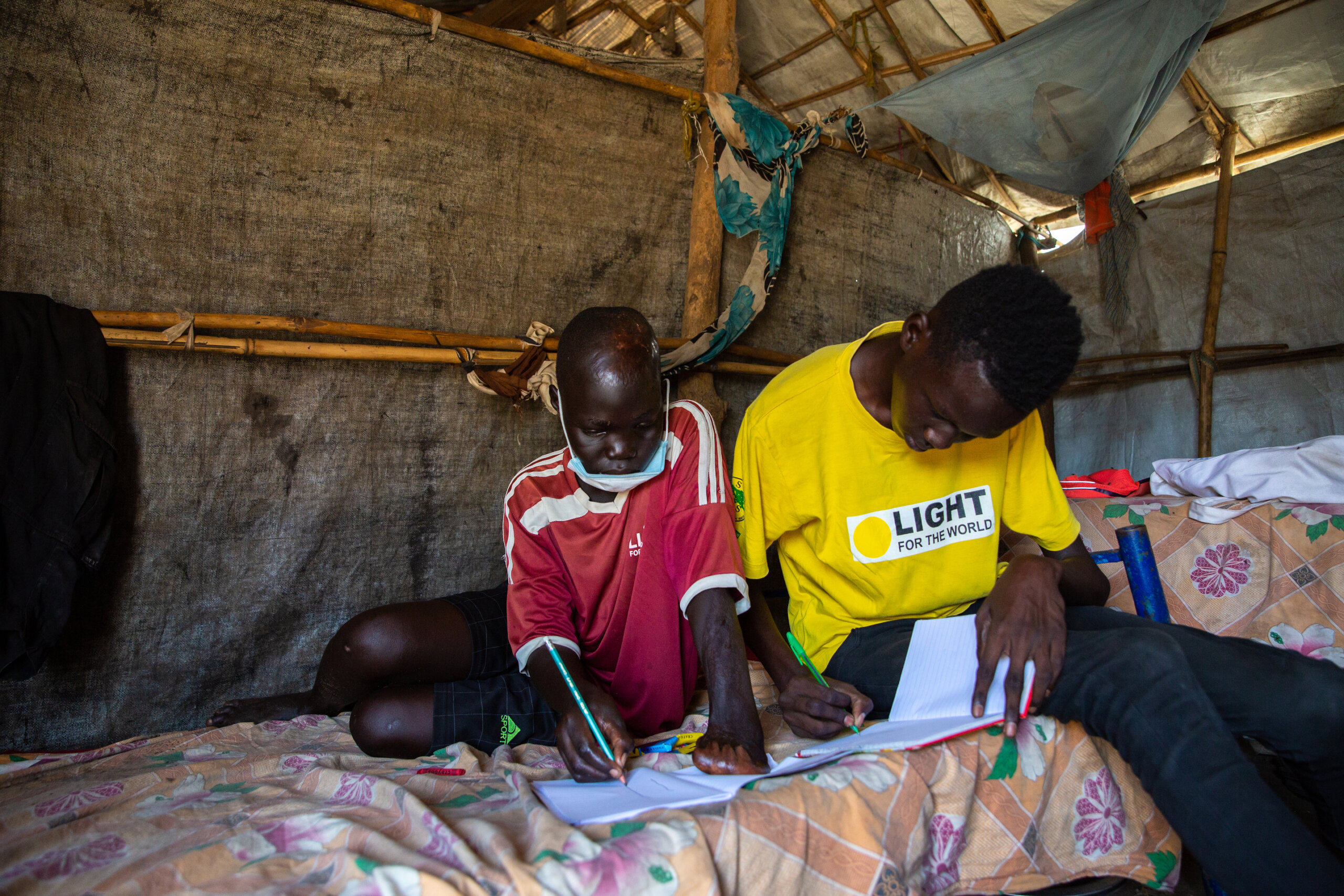 Juma John sits next to his friend and tutor Akoi Mayen Kur in an IDP camp in South Sudan. John is on the left, wearing a red top, while Akoi is wearing a yellow Light for the World top. They both look studious and are writing in books.