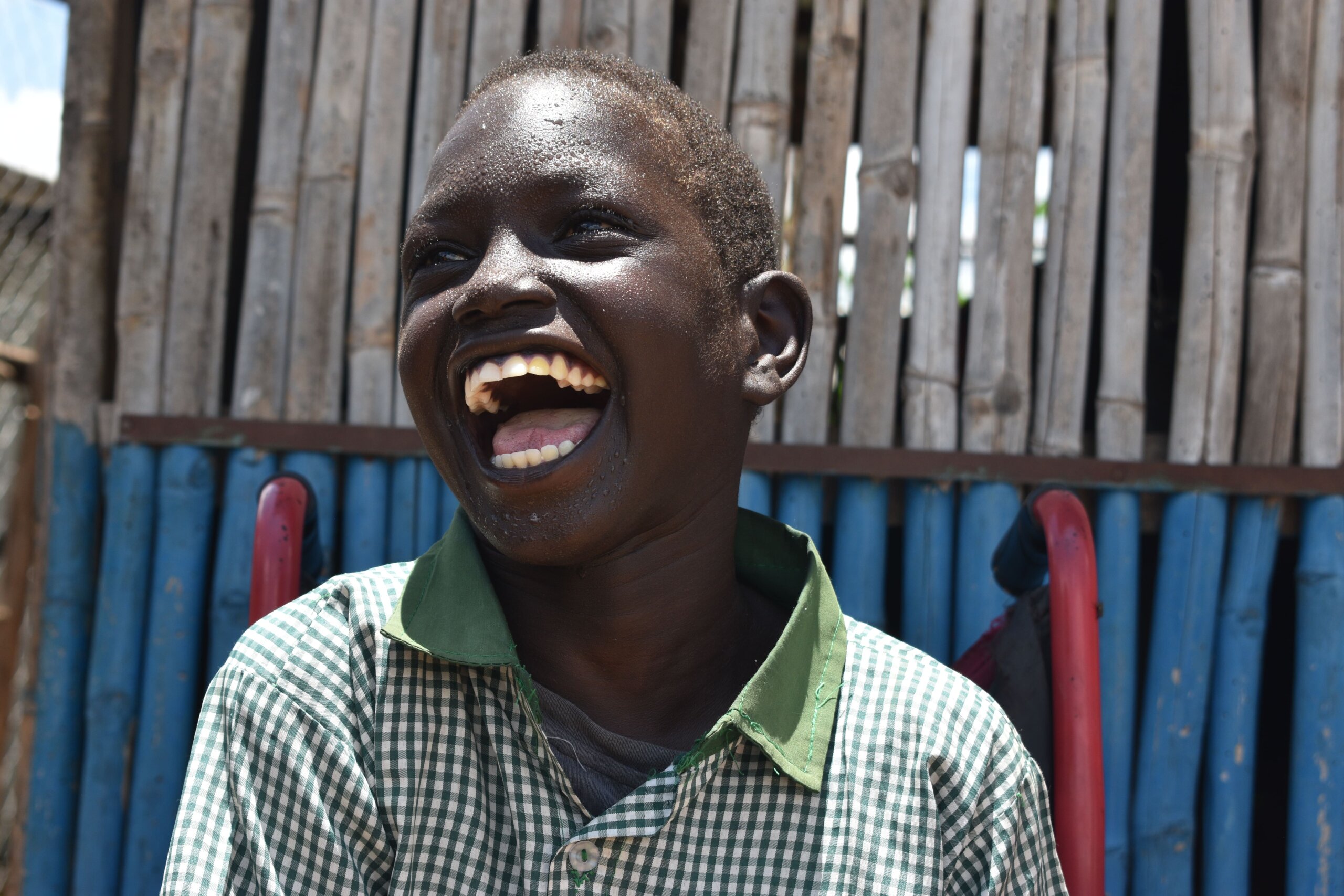 Nyamani Gatluak is pictured smiling outdoors, wearing a checked green shirt, sitting in a wheelchair. There is a wooden building in the background.